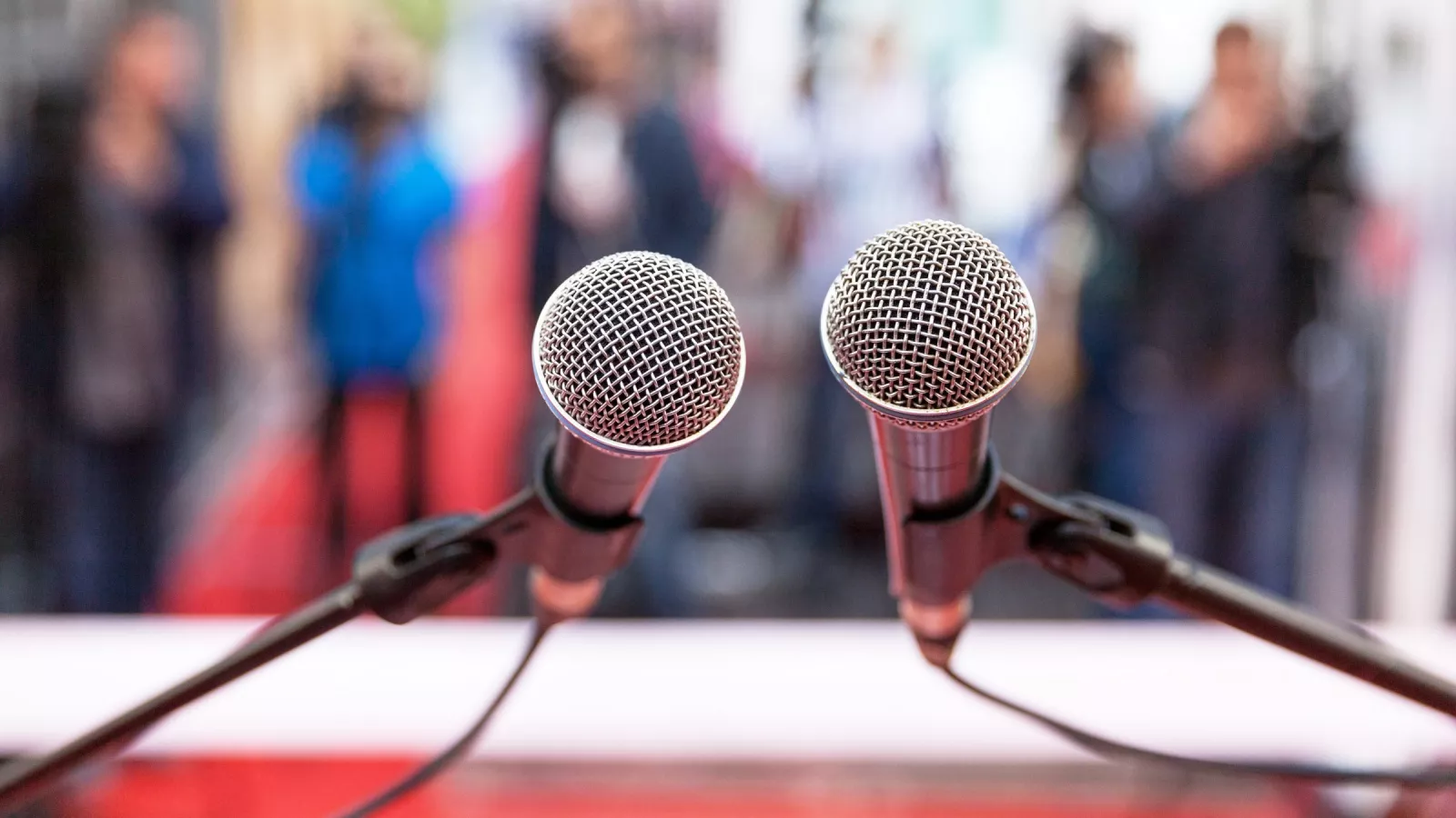 Two microphones attached to microphone stands are crisp and clear in the foreground. In the blurry background, it appears there are several people standing around in suits.