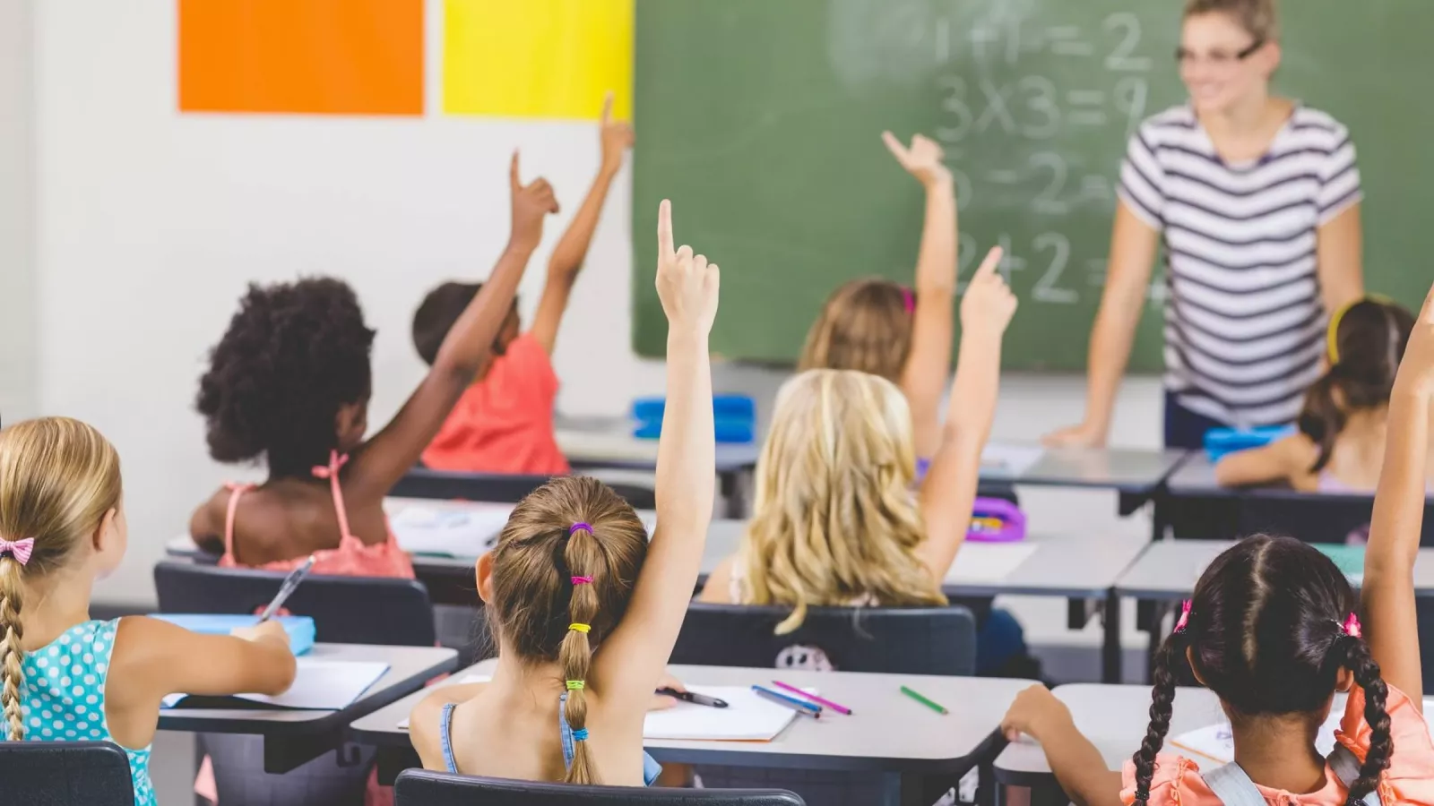 Children sit in desks, hands raised, in front of a smiling teacher in a striped shirt.
