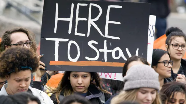 activists protesting immigration policy with sign that says Here to Stay