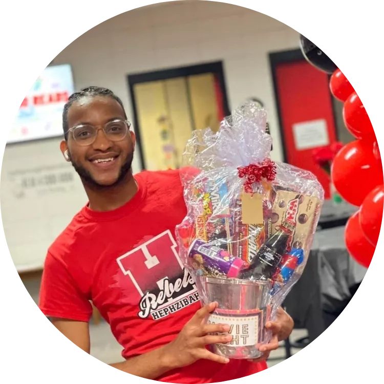 Timothy Lyons smiles for a photo wearing a red school spirit shirt and holding a raffle basket. To his right, there is an arch of black and red balloons.
